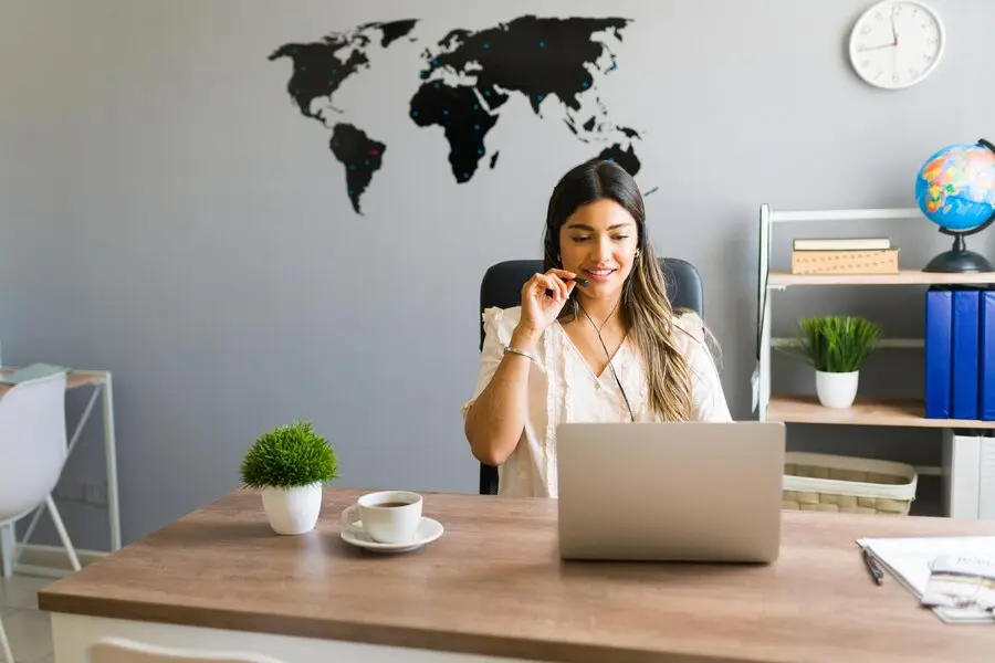 Sur cette image, une femme souriante est assise à un bureau, portant un casque micro, en pleine visioconférence sur son ordinateur portable. Le mur derrière elle est décoré d’une carte du monde noire et d’une horloge, donnant une ambiance internationale. Sur le bureau, une tasse de café, une plante verte et des documents apportent une touche chaleureuse et professionnelle.