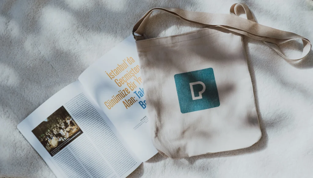 Tote bag en toile beige marqué d'un logo bleu imprimé, posé à plat à côté d'un magazine ouvert